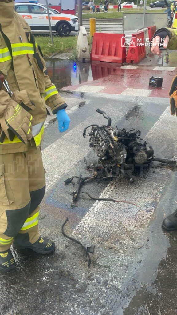 Strażak w mundurze i rękawiczkach niebieskich stoi obok spalonego silnika na mokrej nawierzchni drogi.