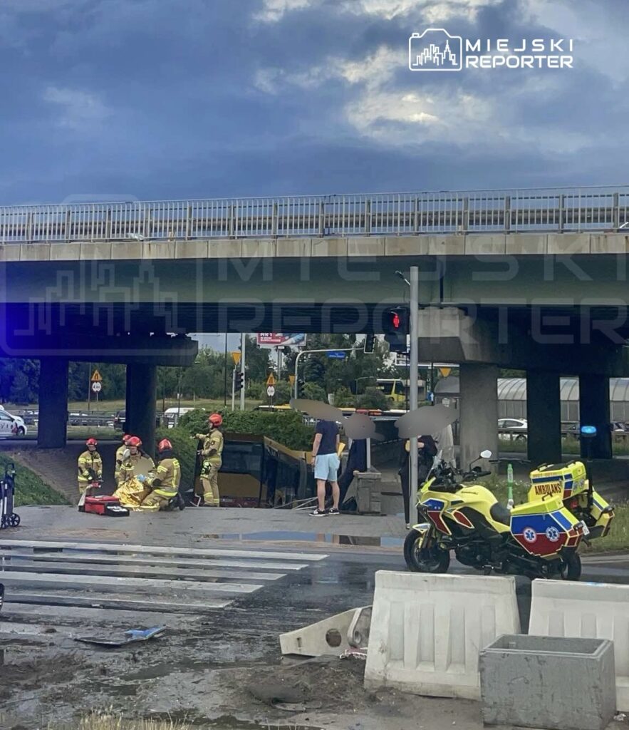 Grupa strażaków w ubraniach ochronnych udziela pomocy poszkodowanemu przy przewróconym pojeździe pod wiaduktem.