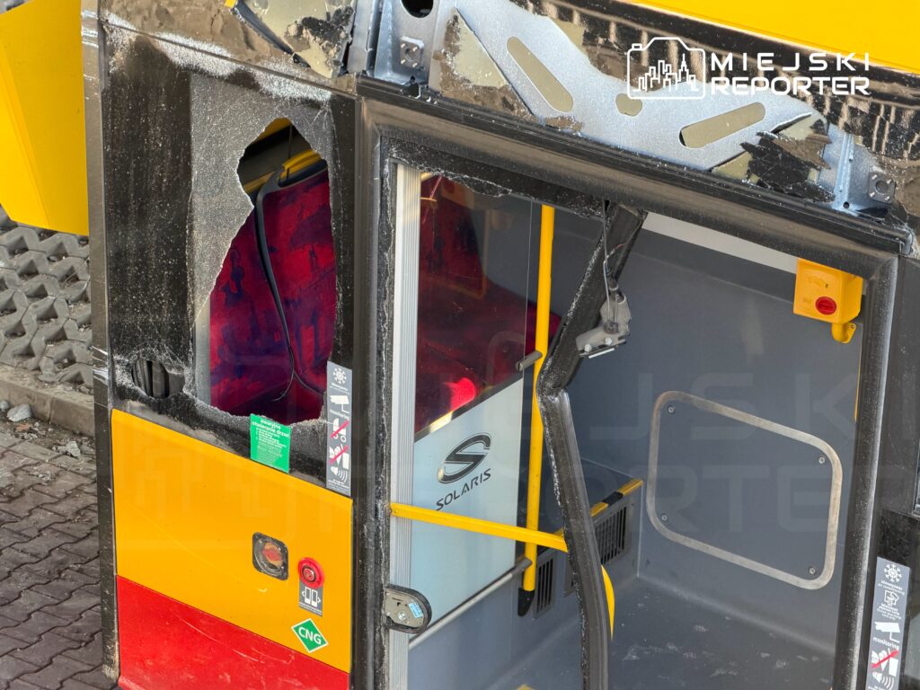 Zniszczona kabina autobusu z wyciętym otworem w drzwiach, widoczne elementy wnętrza i oznaczenia bezpieczeństwa.