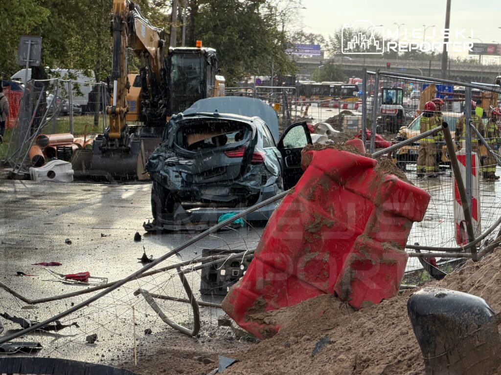 Zniszczony samochód osobowy stoi na zalanej drodze obok sprzętu budowlanego i barier ochronnych.