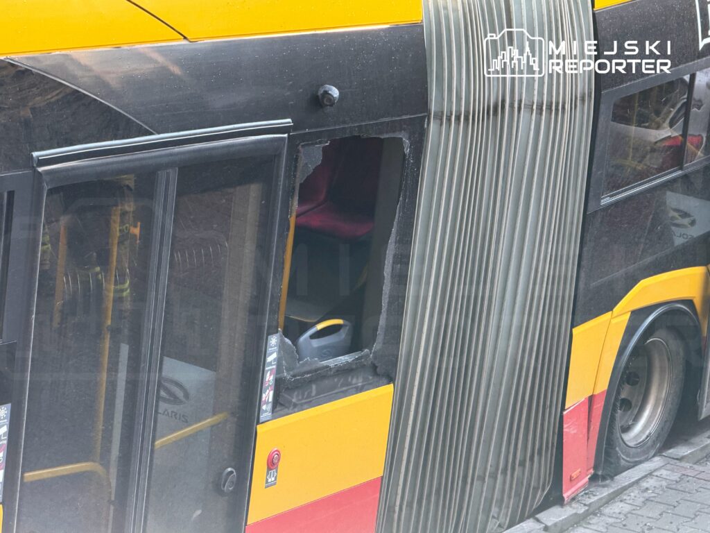 Autobus miejskiego transportu z wybitym oknem, stojący na ulicy w miejskim otoczeniu.