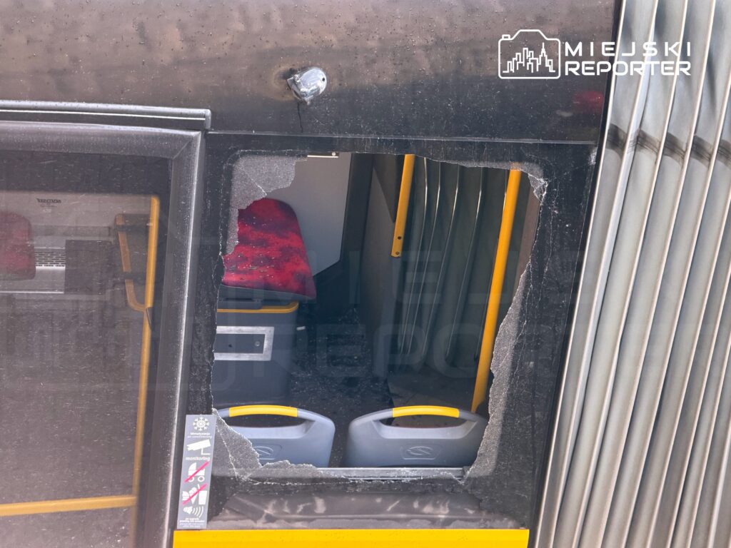 Uszkodzone okno w drzwiach autobusu, przez które widać wnętrze z czerwonymi siedzeniami i żółtymi uchwytami.