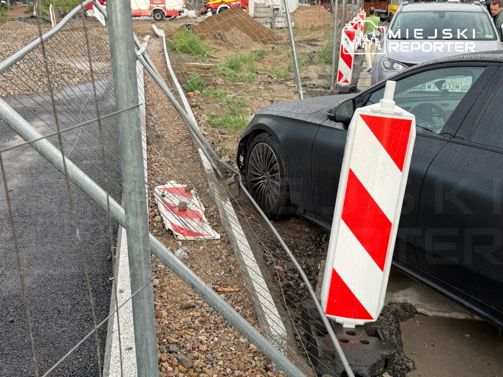 Czarny samochód osobowy wjeżdża w ogrodzenie budowy, a obok znajdują się znaki drogowe i karetka.