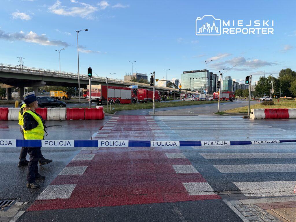 Policjant w żółtej kamizelce zabezpiecza przejście dla pieszych, a w tle widać pojazdy i nowoczesne budynki.