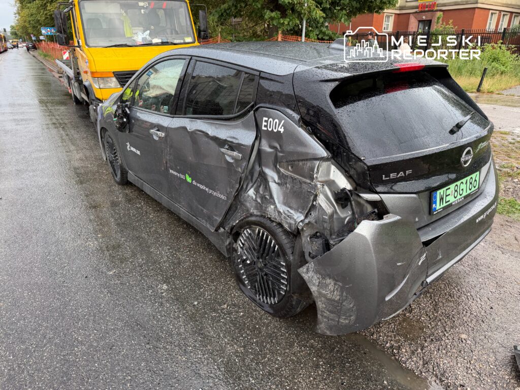 Uszkodzony samochód elektryczny Nissan Leaf z wgniecioną tylną częścią stoi na mokrej nawierzchni obok żółtego pojazdu budowlanego.