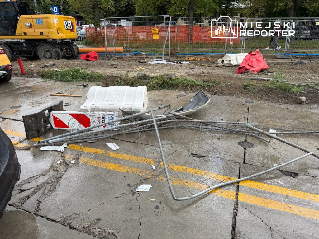 Złomowane elementy infrastruktury leżą na mokrym asfalcie, obok maszyny budowlanej w trakcie prac.