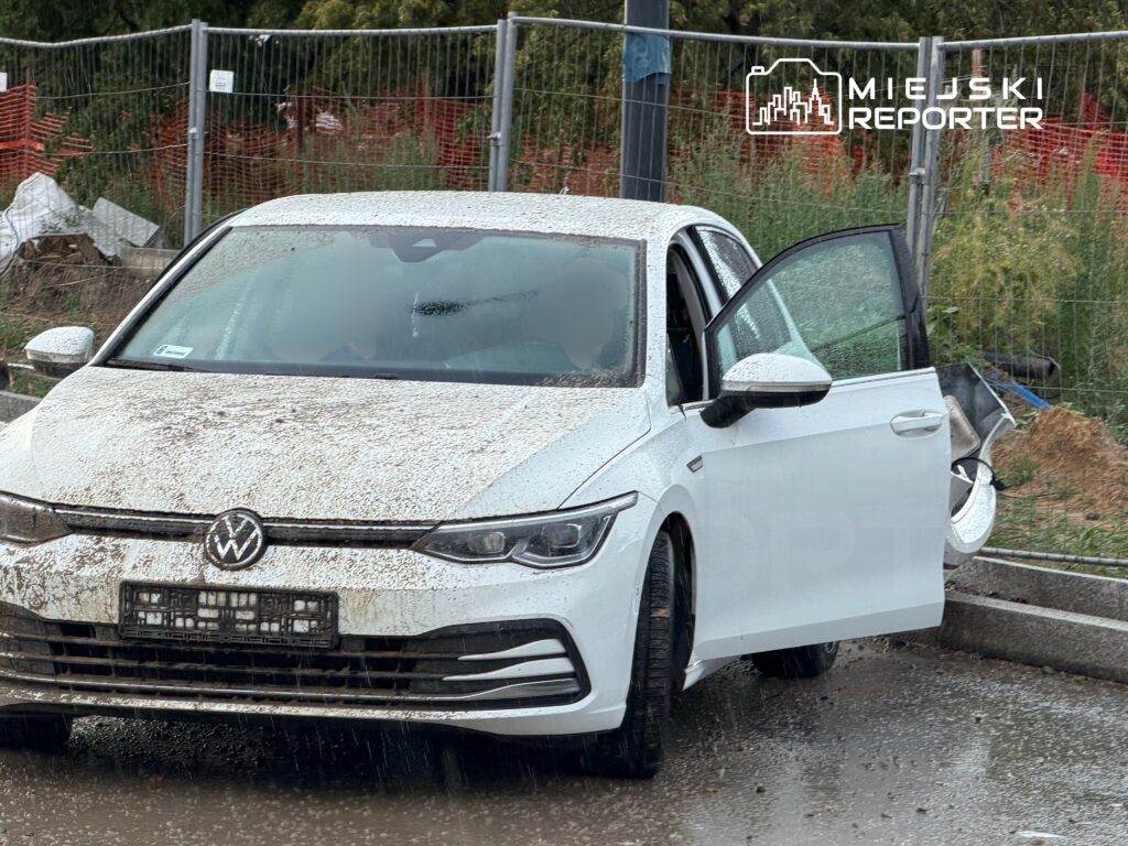 Biały samochód marki Volkswagen z otwartymi drzwiami stoi na mokrej nawierzchni, pokryty błotem. W tle widać ogrodzenie budowy.