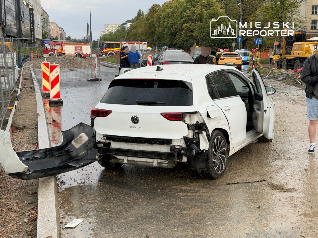 Uszkodzony biały Volkswagen Golf z otwartymi drzwiami stoi na drodze, w tle widoczne służby ratunkowe i roboty drogowe.
