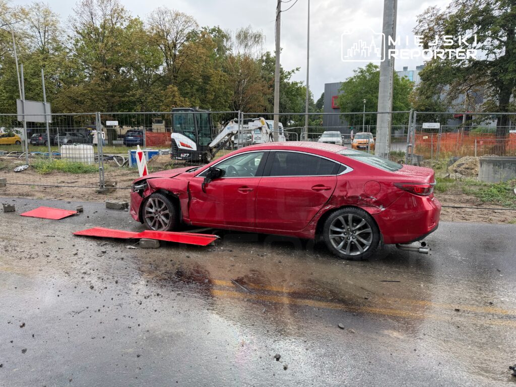 Czerwony samochód osobowy z uszkodzeniem przodu stoi na mokrej nawierzchni, obok placu budowy z ogrodzeniem.