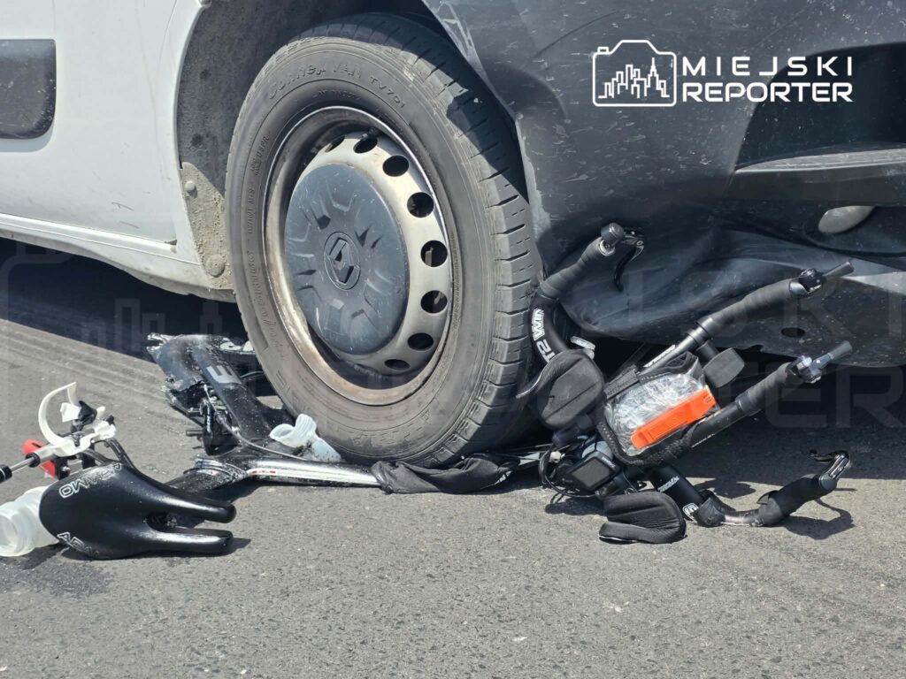 Uszkodzona hulajnoga leży na asfalcie pod samochodem osobowym, jedno z kół pojazdu stoi na jej ramie.