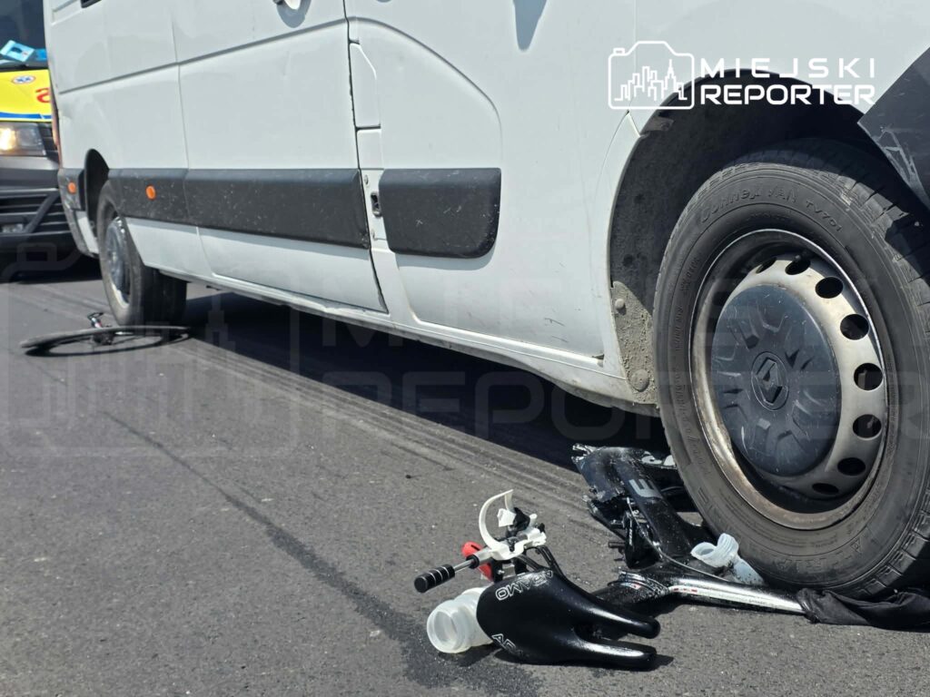Biały furgon z uszkodzoną oponą stojący na drodze, obok leżący sprzęt i fragmenty hulajnogi.