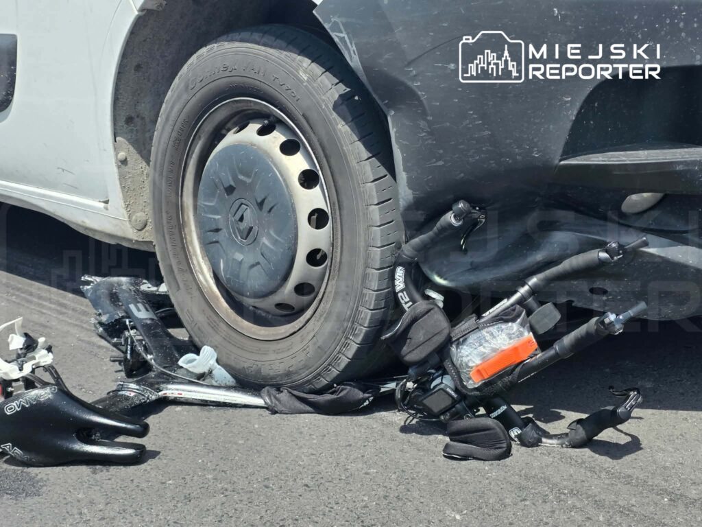 Uszkodzony rower leży pod samochodem, a koło pojazdu znajduje się na jego ramie.