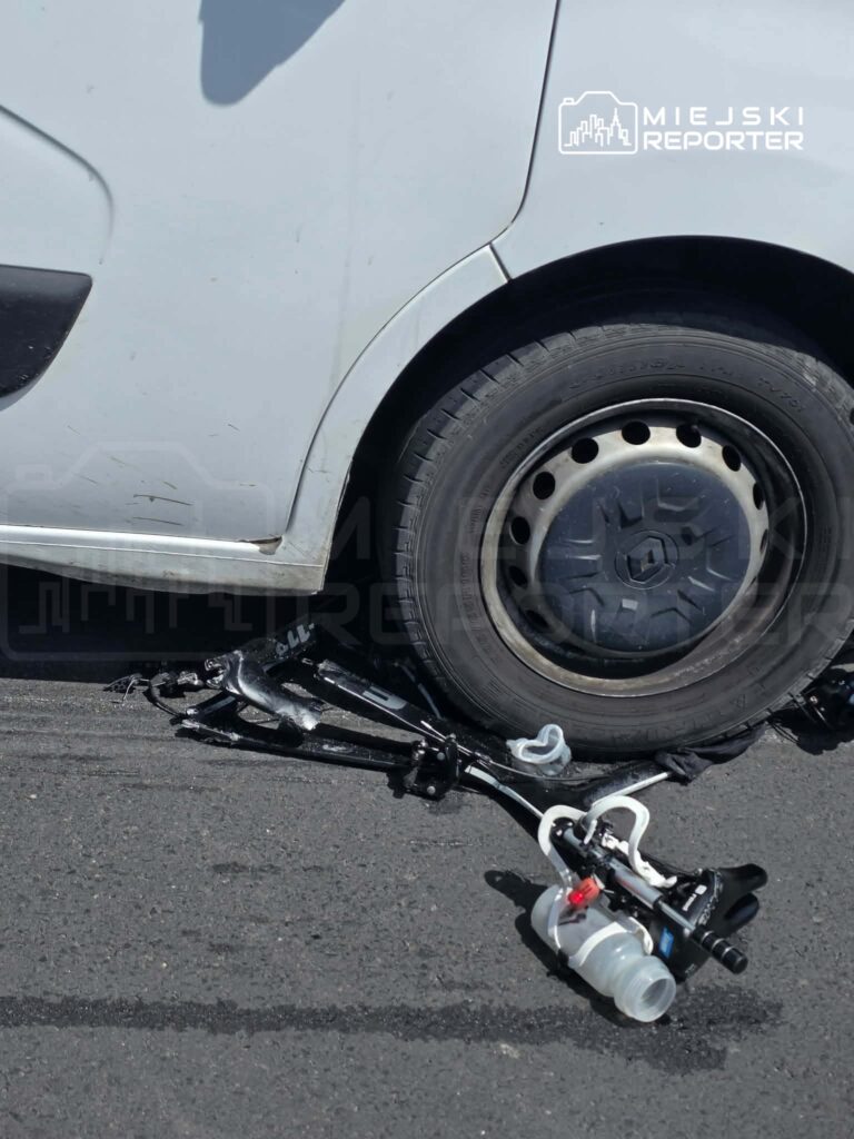 Pojazd z uszkodzoną oponą stojący na asfaltowej nawierzchni, obok leży zniszczona część mechaniczna.