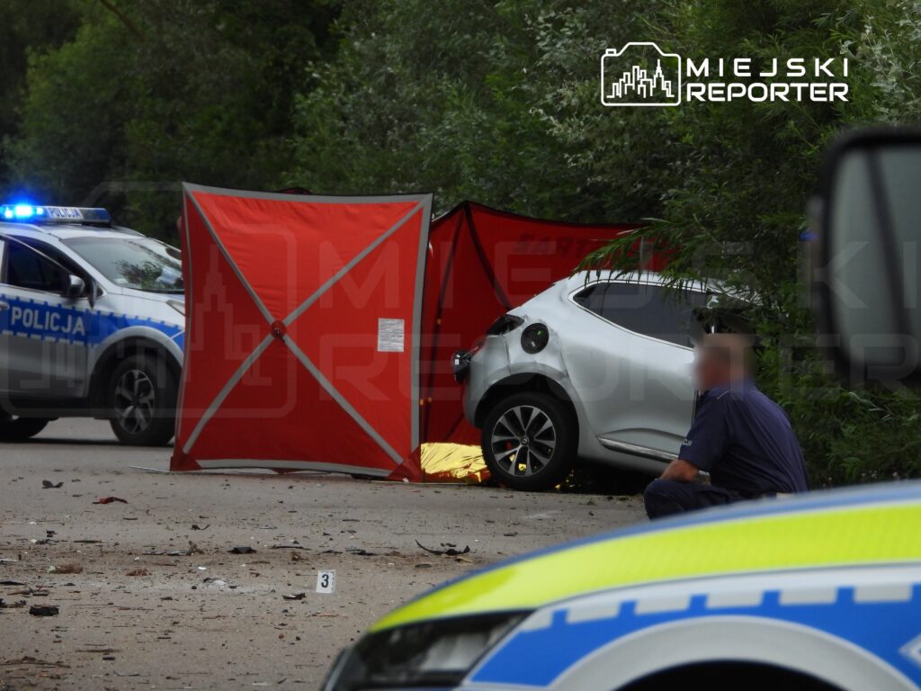 Na drodze stoi policyjny radiowóz, a obok znajduje się rozstawiony czerwony namiot. W tle widać uszkodzone auto oraz funkcjonariusza policji badającego miejsce zdarzenia.