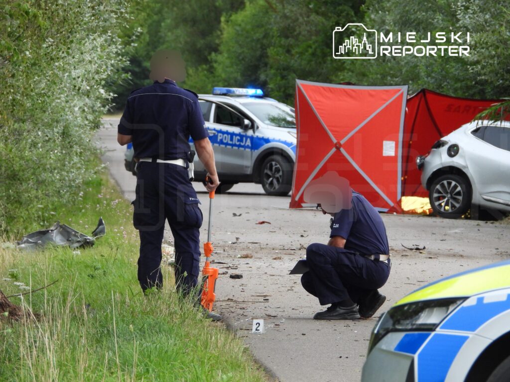 Policjanci analizują miejsce wypadku drogowego na poboczu drogi, obok uszkodzonych pojazdów i oznaczeń.
