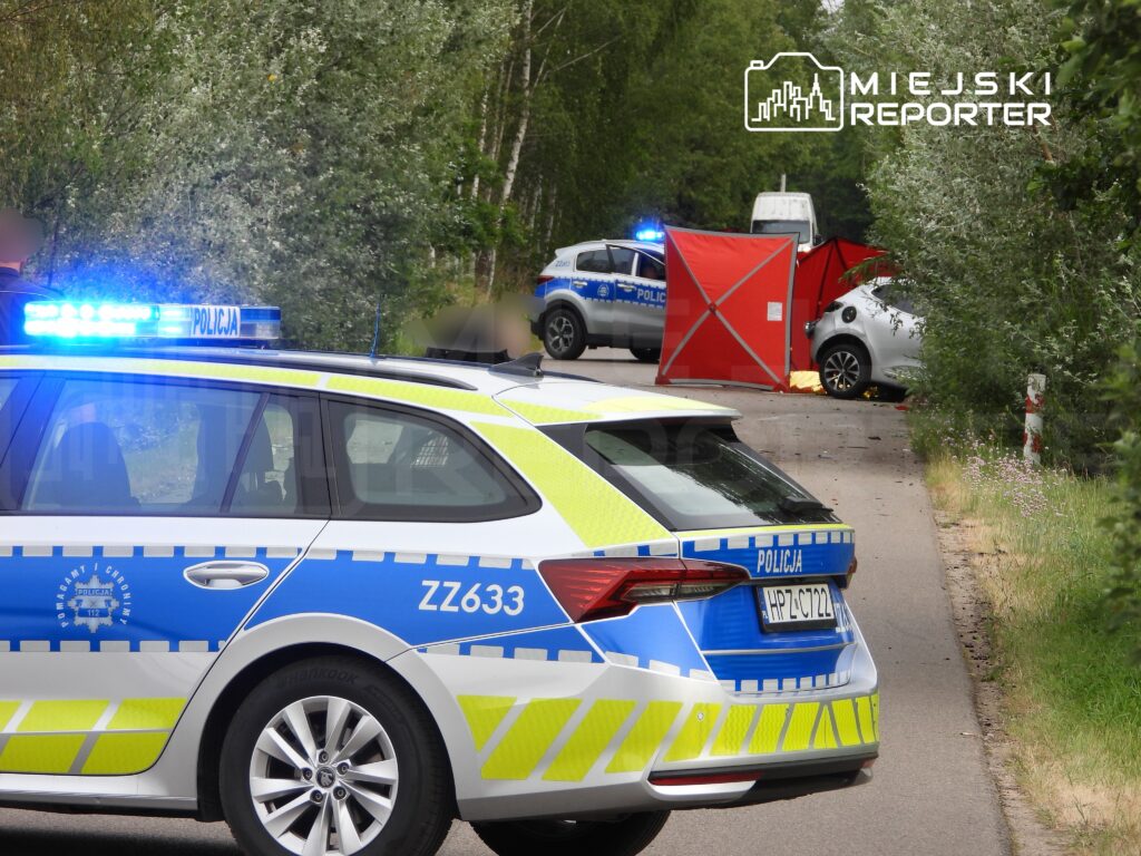 Policyjny radiowóz z włączonymi sygnałami świetlnymi stoi na drodze obok zderzenia dwóch samochodów, w tle widać namiot.