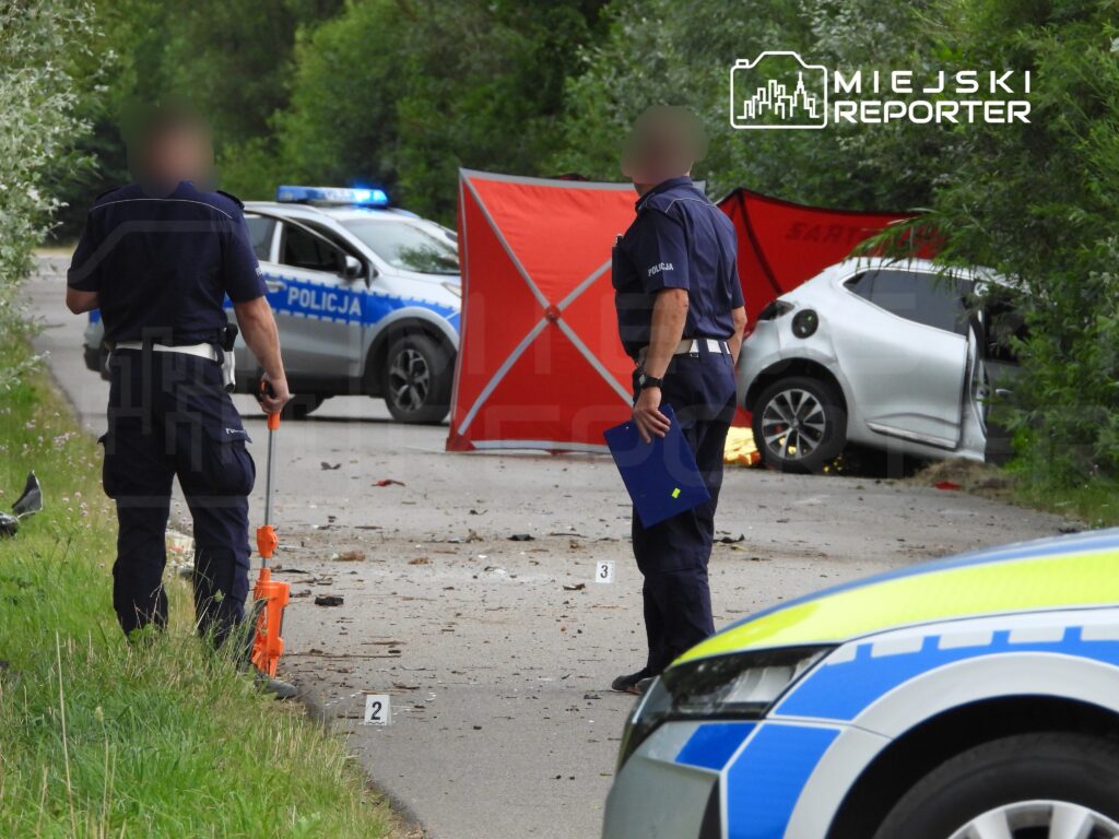 Dwaj policjanci prowadzą dochodzenie na drodze, obok uszkodzonego samochodu i oznaczeń miejsca zdarzenia.