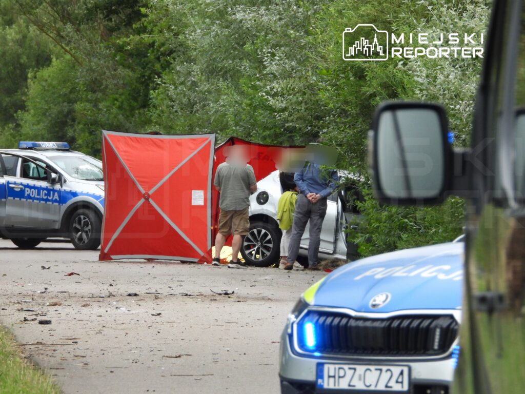 Na drodze stoi mężczyzna w szarej koszuli, rozmawiający z kobietą w zielonej kurtce, obok uszkodzonego samochodu. W tle widoczny radiowóz policyjny.