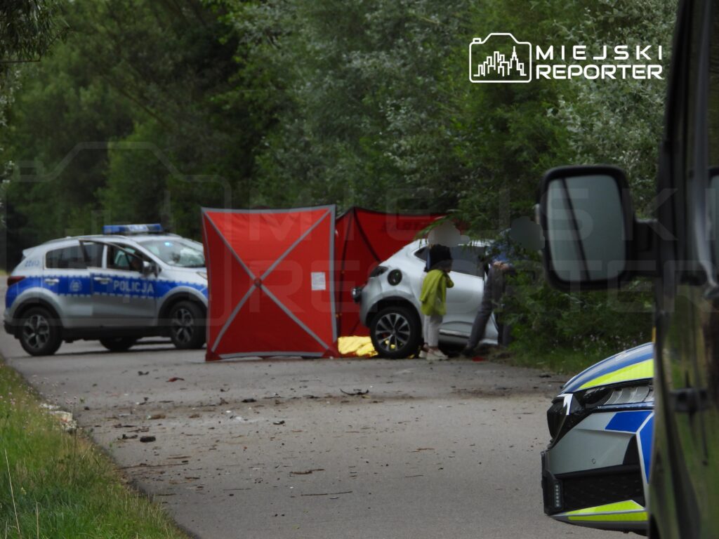 Na drodze stoi czerwona osłona, a obok białego samochodu rozmawia kilka osób, w tym jedna w zielonej kurtce. W tle widać policyjne auta.
