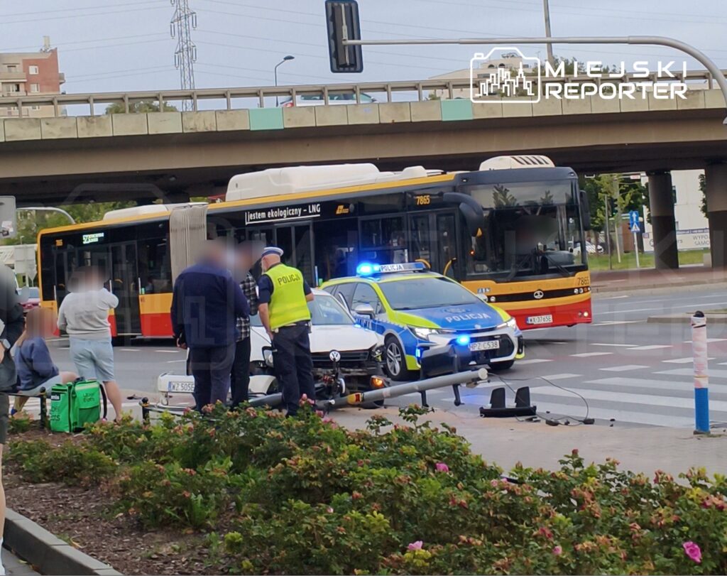 Grupa osób obserwująca policyjną interwencję przy wypadku drogowym, obok uszkodzonego samochodu i autobusu.