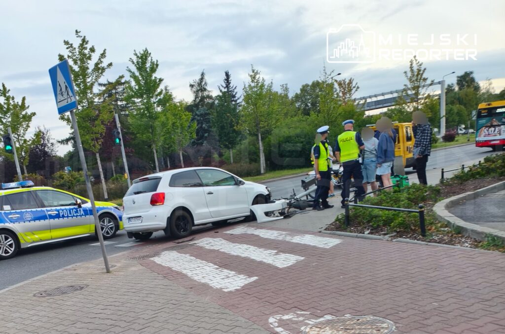 Biały samochód osobowy stoi na chodniku obok grupy osób oraz policjantów w kamizelkach odblaskowych.
