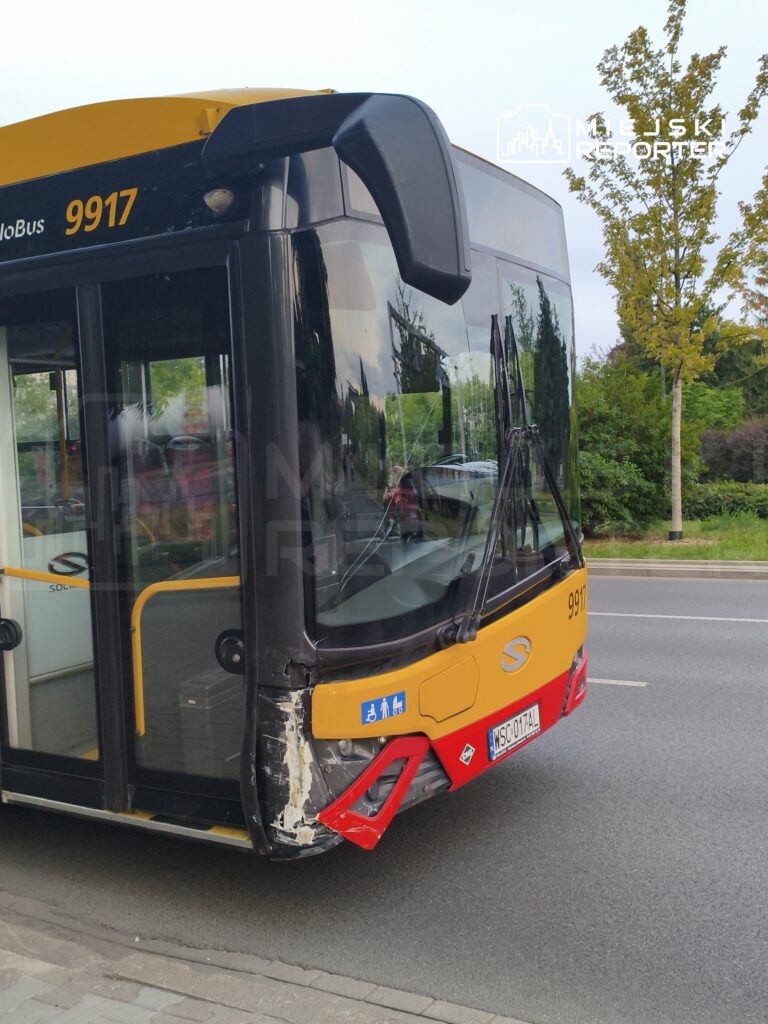 Autobus miejski z uszkodzeniem przedniego zderzaka stoi na ulicy, obok rosnące drzewa i trawniki.