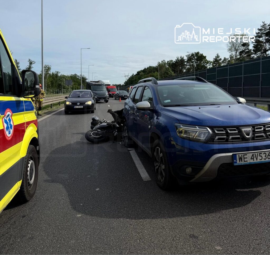 Motocykl leży na jezdni obok niebieskiego SUV-a, a w tle widać stojące samochody i karetkę.