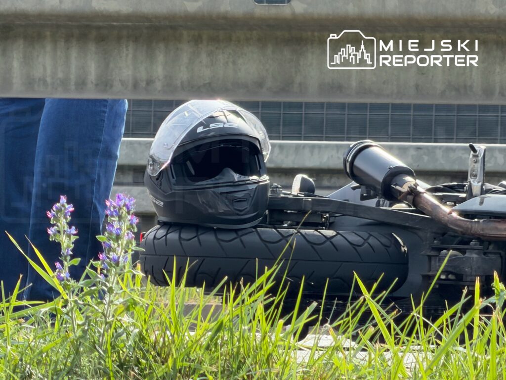 Czarny kask motocyklowy leży na oponie, obok widoczne są zielone trawy i kwiaty. W tle stoi osoba w niebieskich dżinsach.