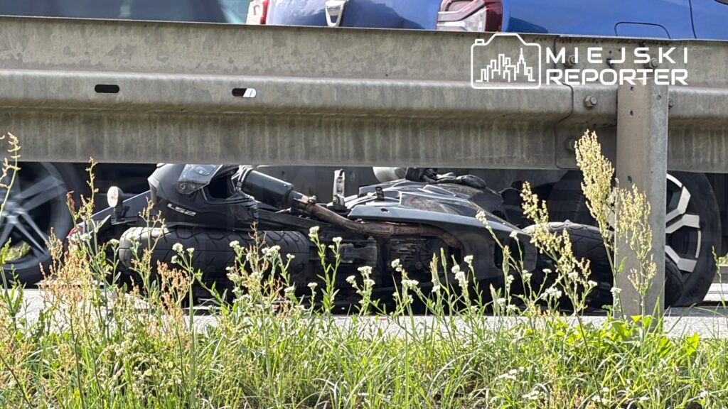 Motocykl leżący na poboczu drogi, otoczony wysoką trawą i kwiatami, obok ruchu samochodowego.
