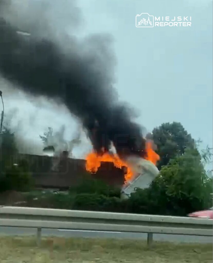 Pojazd w płomieniach, wydobywający gęsty czarny dym, znajduje się obok drogi, w otoczeniu zieleni.
