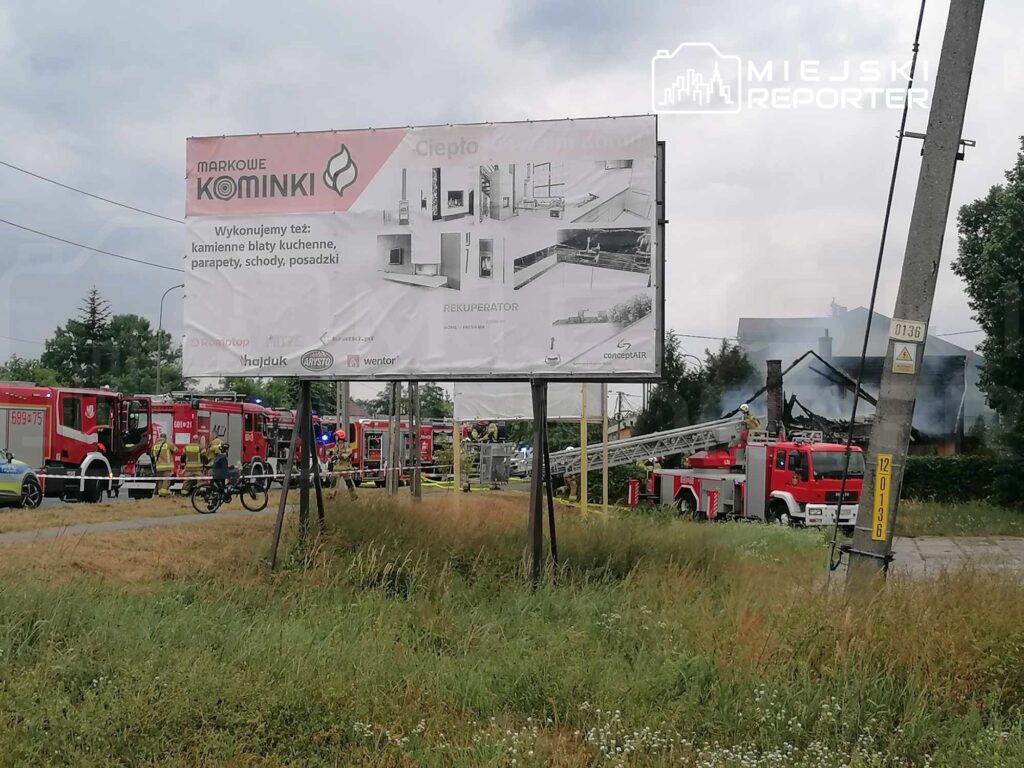 Na drodze widoczne są strażackie pojazdy, a w tle unosi się dym z palącego się budynku. W pobliżu stoi billboard reklamowy.