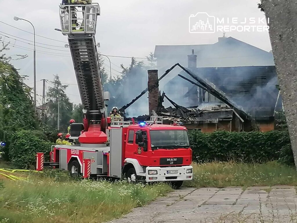 Strażacy na wozie strażackim z drabiną gaszą pożar zniszczonego budynku, z którego unosi się dym.
