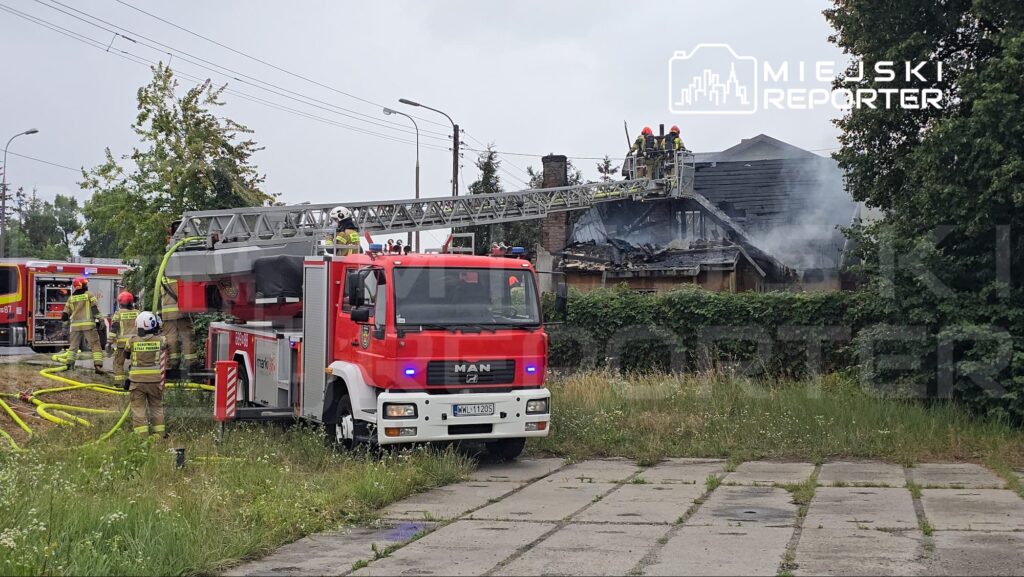 Strażacy pracują nad gaszeniem pożaru na dachu budynku, korzystając z węża pożarniczego z wozu strażackiego.