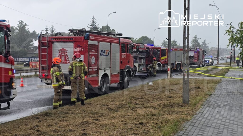Grupa strażaków w mundurach omawia sytuację obok wozów strażackich na deszczowej ulicy.