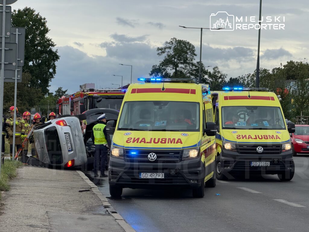 Na drodze stoi przewrócony samochód, obok pracują strażacy i policjant, a w tle widać karetki pogotowia.