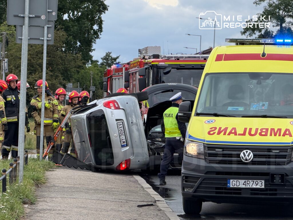 Grupa strażaków w hełmach obserwuje przewrócone na bok auto, obok stoi karetka pogotowia i radiowóz.