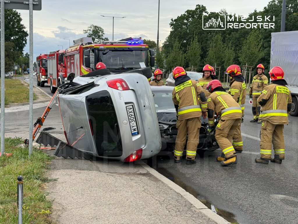 Grupa strażaków w mundurach ratunkowych podnosi przewrócone auto na drodze, obok widoczne wozy strażackie.
