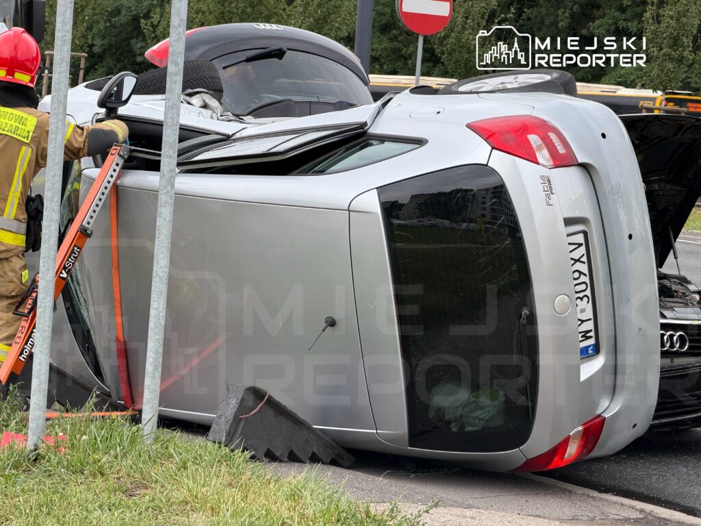 Strażak w hełmie, używający narzędzi do podnoszenia przewróconego samochodu na poboczu drogi.