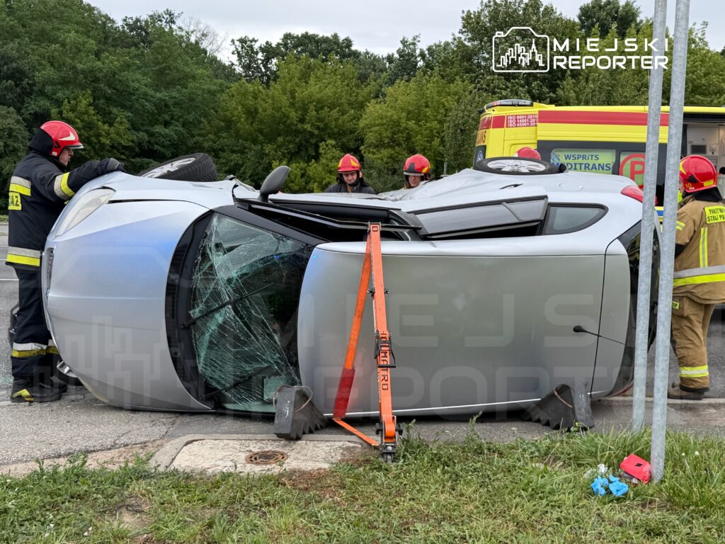 Strażacy zabezpieczają przewrócone na bok auto osobowe przy drodze, obok widoczna karetka pogotowia.
