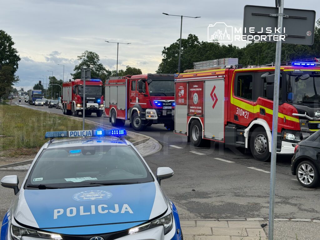 Cztery wozy strażackie oraz radiowóz policyjny zatrzymane na drodze, w tle ruch samochodowy.