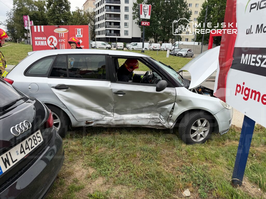 Kierowca w uszkodzonym samochodzie osobowym stoi obok pojazdu na terenie parkingu, obok znaku reklamowego KFC.