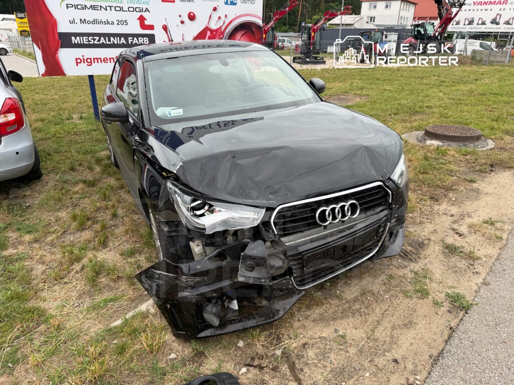 Czarny samochód marki Audi z uszkodzonym przodem stoi na trawie obok drogi, w tle widoczny billboard.