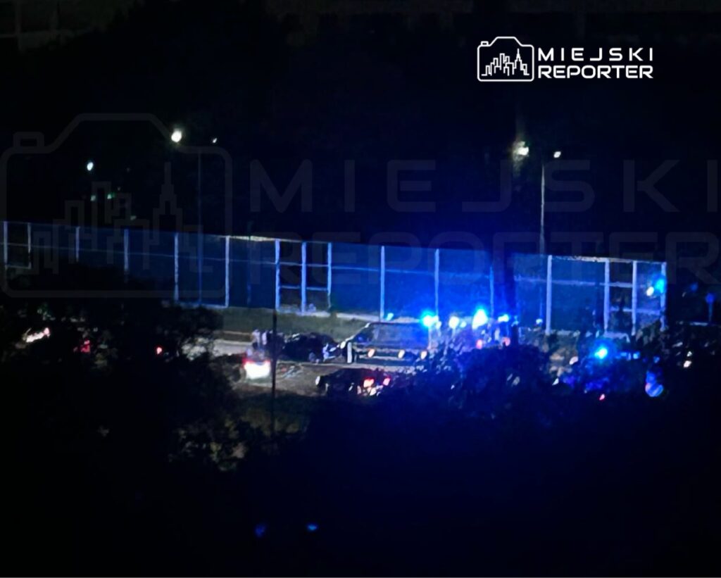 Grupa policjantów z niebieskimi sygnałami świetlnymi zabezpiecza teren przy ogrodzeniu boiska w nocy.
