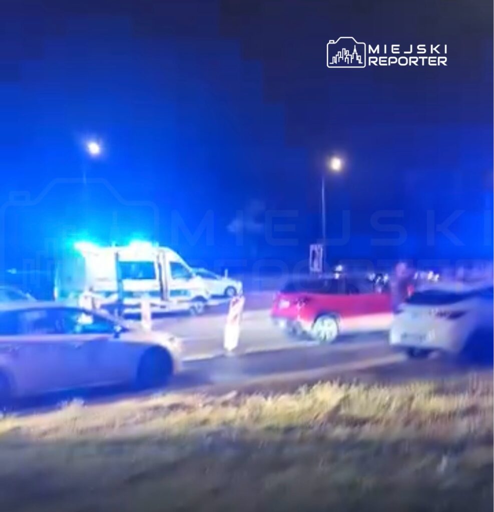 W nocy na drodze zatrzymane są samochody, a w tle widać niebieskie światła radiowozu i karetki.