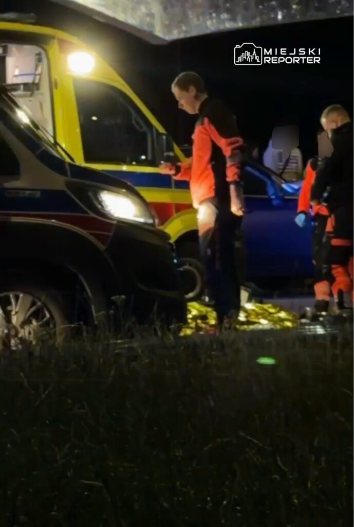 Ratownicy medyczni w pomarańczowych strojach pracują przy ambulansie na oświetlonym poboczu drogi w nocy.