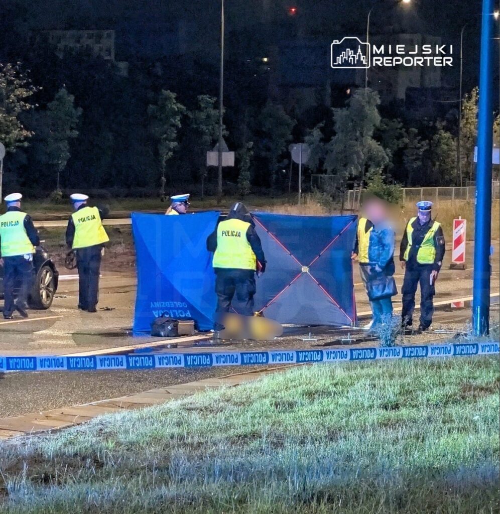 Grupa policjantów zabezpiecza miejsce zdarzenia, stojąc obok niebieskiej bariery na oświetlonej ulicy nocą.