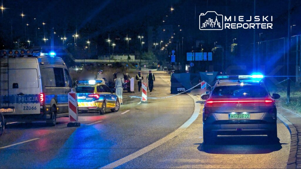 Policjanci zabezpieczają miejsce zdarzenia na drodze, przy włączonych sygnałach świetlnych radiowozów.