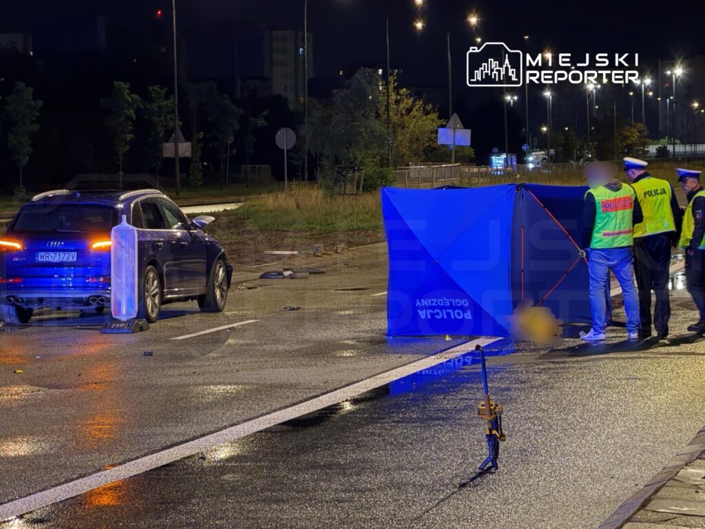Na jezdni, obok niebieskiego SUV-a, policjanci prowadzą czynności dochodzeniowe przy namiocie zabezpieczającym.