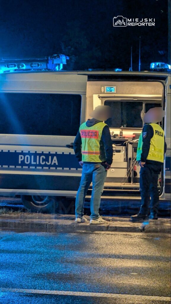 Dwaj policjanci w odblaskowych kamizelkach rozmawiają obok radiowozu w oświetlonym niebieskim blasku.