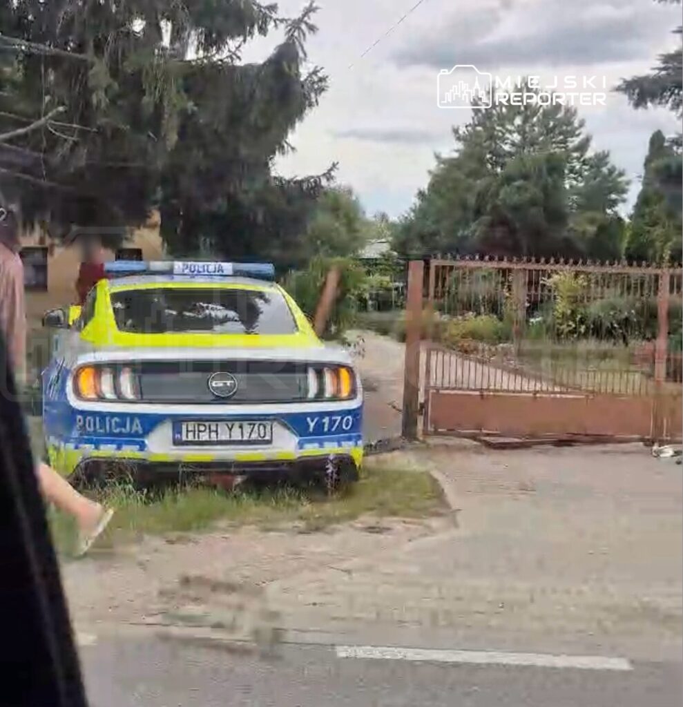 Policjant obok radiowozu z niebiesko-żółtym oznakowaniem na posesji z ogrodzeniem.
